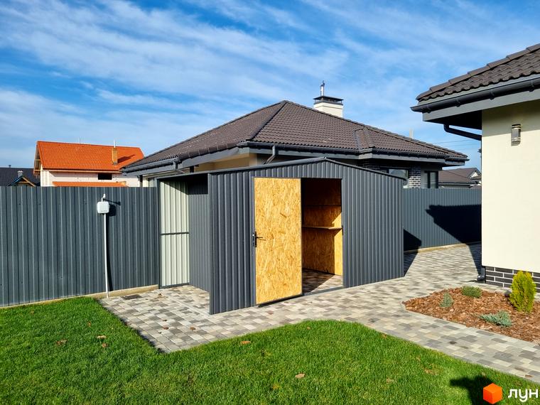 Котеджне містечко КМ Сонячний: сіра металева господарська споруда з дерев'яними дверями та полицями всередині, розташована на вимощеній плиткою ділянці поруч із зеленим газоном та парканом.