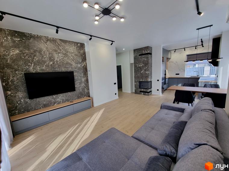 Інтер'єр вітальні з каміном та кухні в житловому комплексі КМ Сонячний. Простора вітальня з телевізором на мармуровій стіні та сірим диваном. Сучасна кухня з обіднім столом.