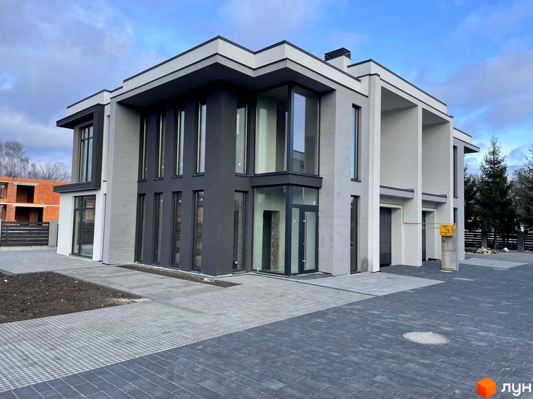 Сучасний двоповерховий будинок з великими вікнами та сірою обробкою, розташований на бруківці з доглянутою ділянкою.