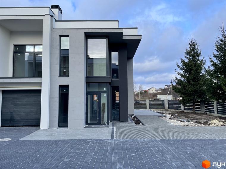 Сучасний дуплекс HEG WOW HOUSE з великими вікнами та гаражем. Перед будинком вимощена сіра плитка та зелені ялинки.