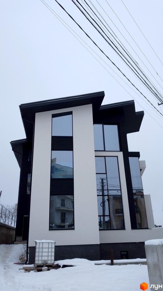 Сучасна новобудова з панорамними вікнами та чорно-білим фасадом на вулиці Урожайна. Навколо будинку засніжена територія та будівельні матеріали.