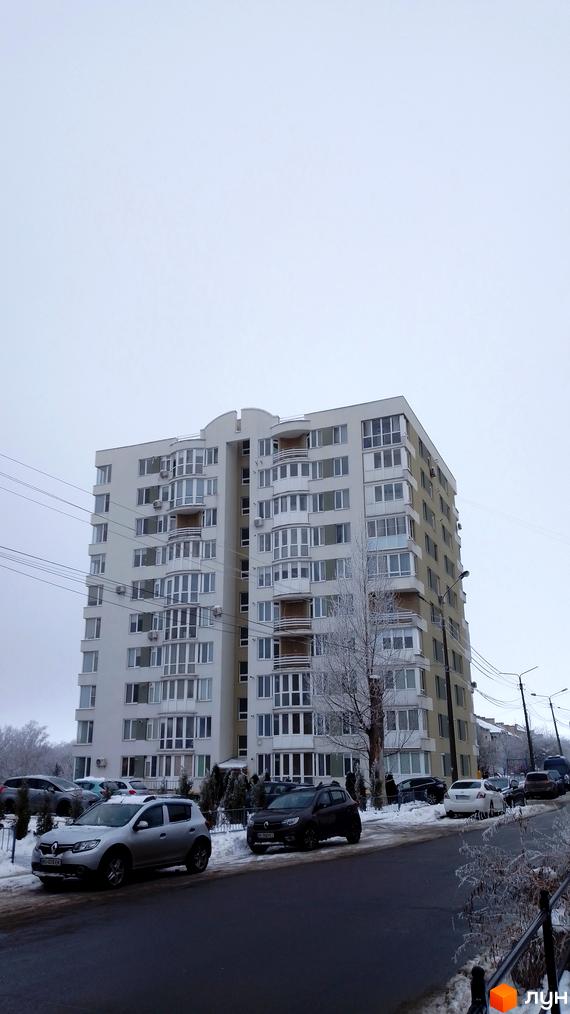 Сучасна новобудова на вулиці Золотогірській у Києві. Будинок має білий фасад з численними заскленими балконами та кондиціонерами. Навколо будинку розташована прибудинкова територія з деревами та припаркованими автомобілями.