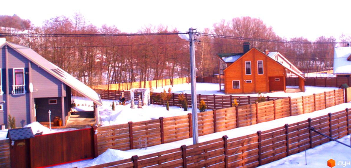 Зовнішній вигляд котеджного містечка КМ Заріччя з дерев'яними будинками та прибудинковою територією, огородженою парканом.