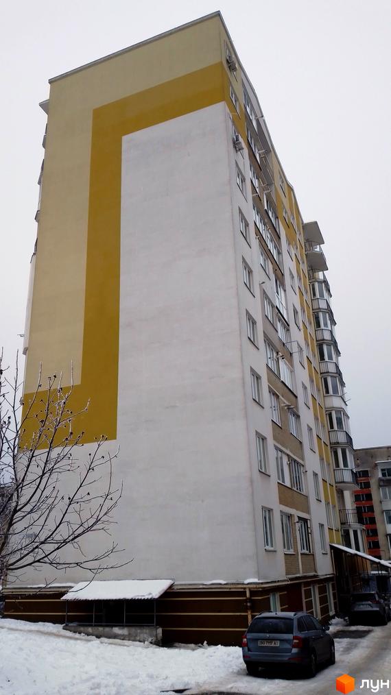 Зовнішній вигляд житлового комплексу на вул. Золотогірська, 14а, з жовто-білим фасадом та балконами. Прибудинкова територія вкрита снігом, стоять автомобілі.