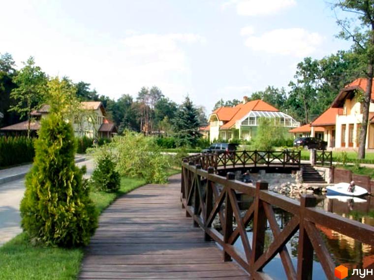 Комплекс КМ Bosco Village з котеджами та доглянутою прибудинковою територією, пішохідна доріжка та місток над водоймою.