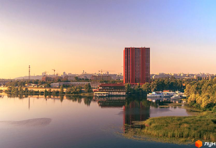 Житловий комплекс DOCK32 – висотна будівля червоного кольору на березі річки з причалом та міським пейзажем на горизонті.