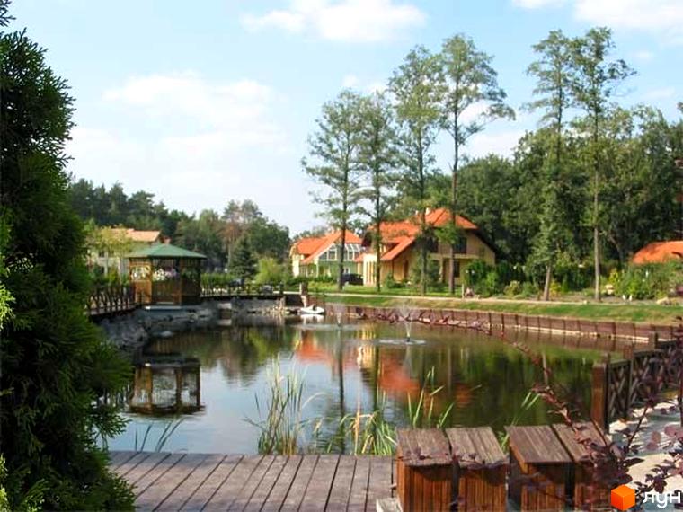 Житловий комплекс Bosco Village. Мальовничий будинок з помаранчевим дахом біля озера. На території містечка котеджів є місток, альтанка та зелені насадження.