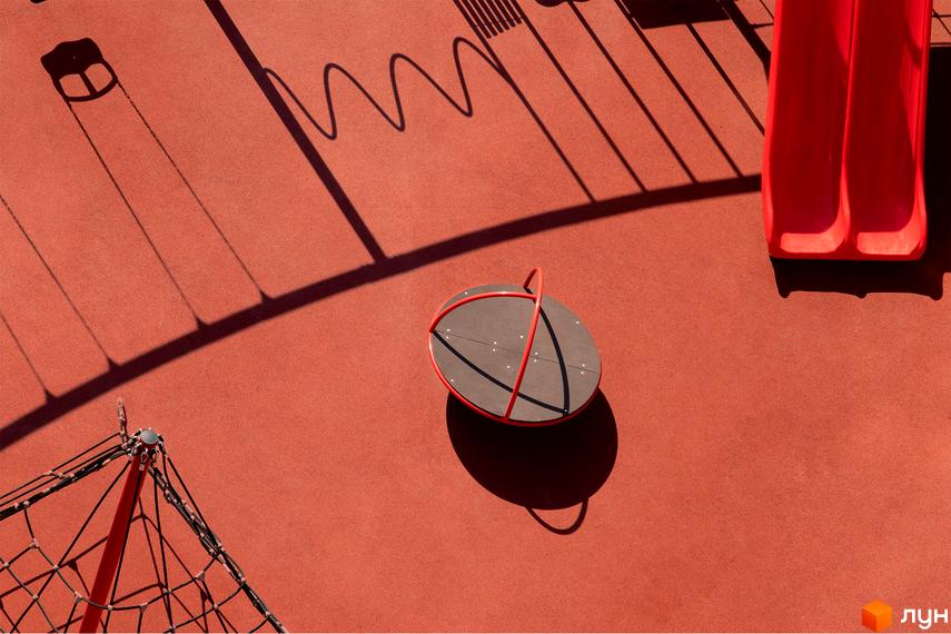 Ігровий майданчик з червоним покриттям та тінями від гойдалок і гірки на території житлового комплексу DOCK32.