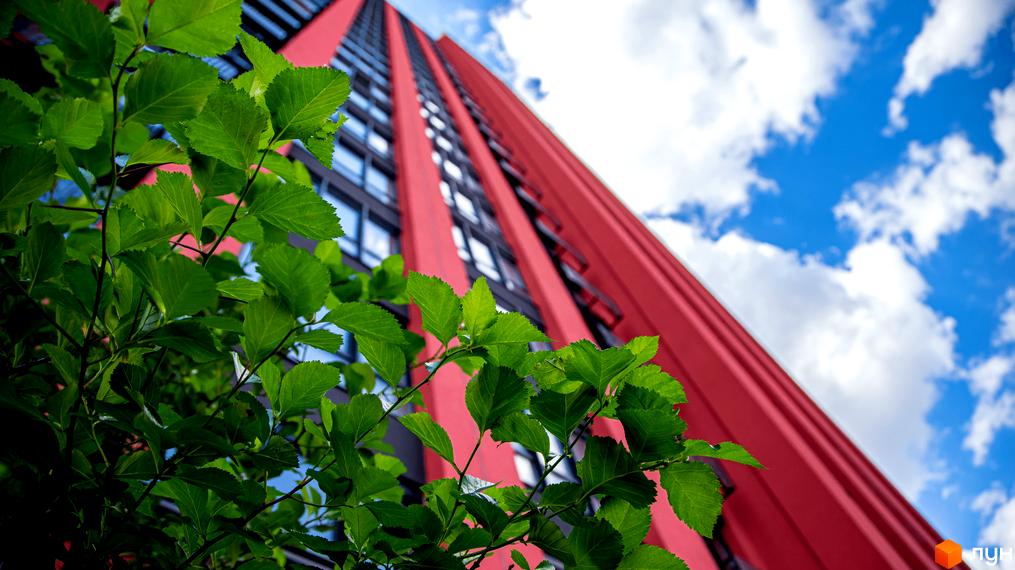Високий червоний будинок житлового комплексу DOCK32 з великими вікнами, що видніється крізь зелене листя на тлі блакитного неба.