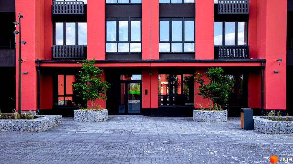 Червоний фасад житлового комплексу DOCK32 з чорними вікнами та балконами. Прибудинкова територія викладена бруківкою, озеленена деревами у габіонних клумбах.