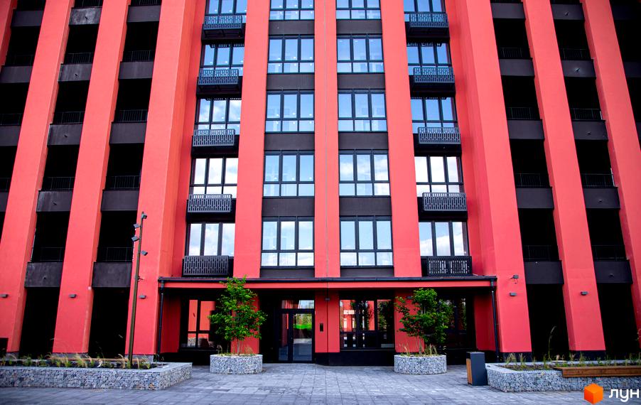 Фасад житлового комплексу DOCK32 виконаний у червоно-чорних кольорах з великими вікнами. Прибудинкова територія облаштована бруківкою та зеленими насадженнями.