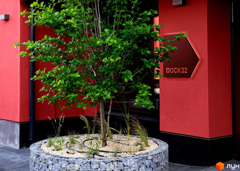 Фасад житлового комплексу DOCK32 виконаний у червоному кольорі з чорною основою. Перед будинком розташована кругла клумба з деревом та системою поливу.
