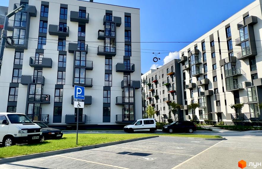 Житловий комплекс Проект Панорама з білими фасадами та балконами, парковка з розміткою для авто. На території є газон та зелені насадження.