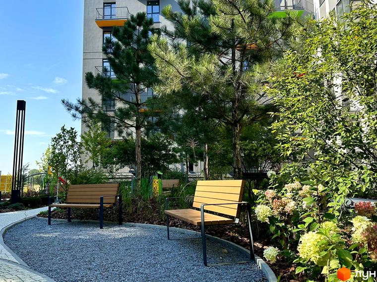 Житловий комплекс Проект Панорама з фасадом сірого кольору, балконами з помаранчевими вставками. На прибудинковій території лавки, дерева та квіти.