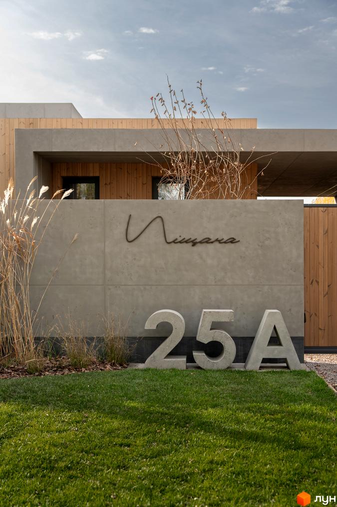 Фасад будинку з написом «Піщана» та номером 25А. Навколо зелена трава та рослини, що створюють атмосферу спокою.