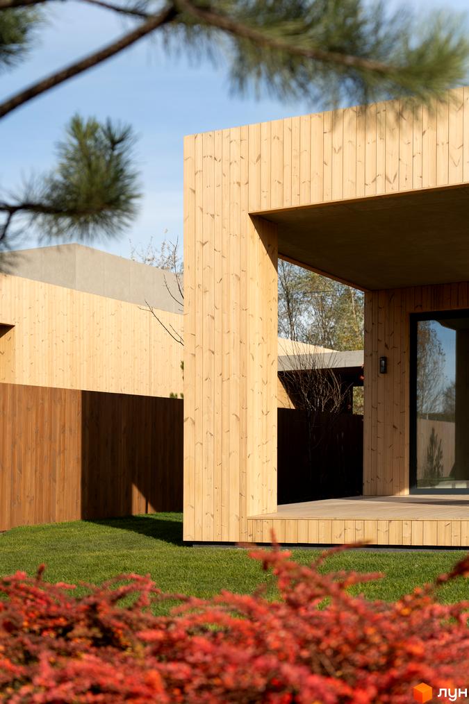 Житловий комплекс з дерев'яним фасадом на тлі зеленої трави та рослин. На задньому плані видно огорожу та частину іншого будинку.