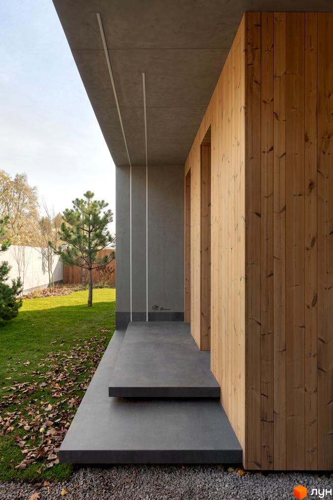 Сучасний вхід до будинку з дерев'яним оздобленням та бетонними сходами. Навколо зелений газон та дерева.