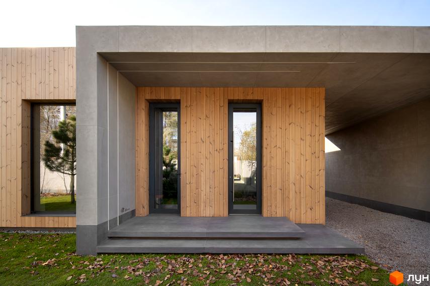 Фасад будинку з дерев'яним оздобленням та великими вікнами. На передньому плані газон з опалим листям, поруч доріжка з гравію.