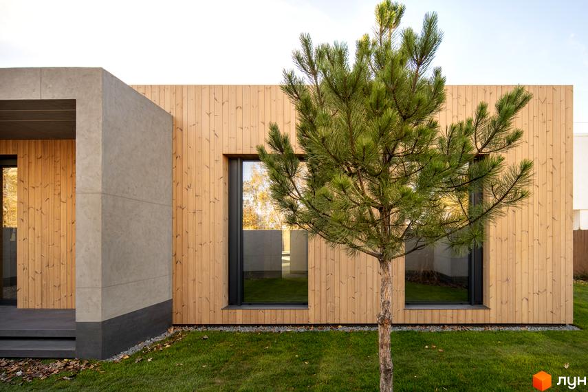 Зовнішній вигляд будинку з дерев'яним фасадом та великими вікнами. На передньому плані зелена трава та дерево, що росте на прибудинковій території.