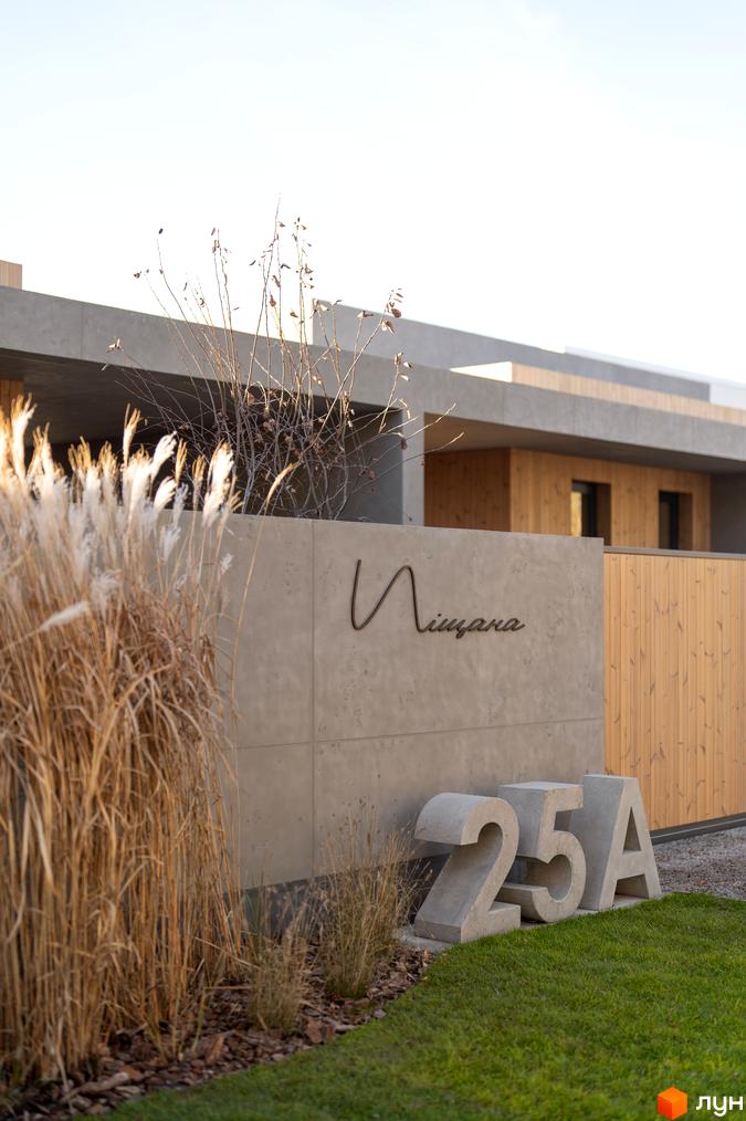Фасад житлового комплексу на вулиці Піщана, 25а, 25б, з бетонною стіною та об'ємними цифрами 25А. Прибудинкова територія з травою та декоративними рослинами.
