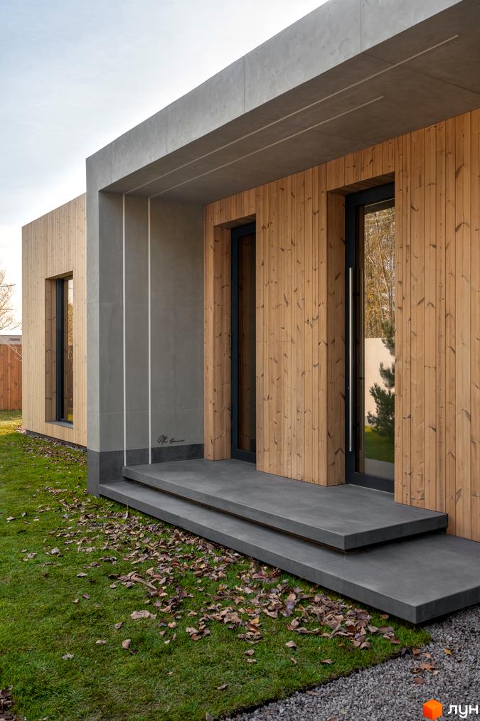 Сучасний житловий комплекс на вулиці Піщаній, 25а, 25б, з дерев'яним фасадом та бетонними елементами. Навколо будинку зелена галявина з опалим листям.