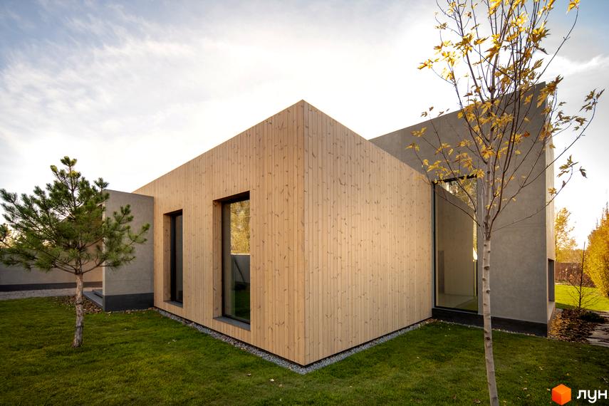 Сучасний будинок з дерев'яним фасадом та сірими бетонними елементами на вулиці Піщана, 25а, 25б. Навколо будинку зелений газон та молоді дерева.