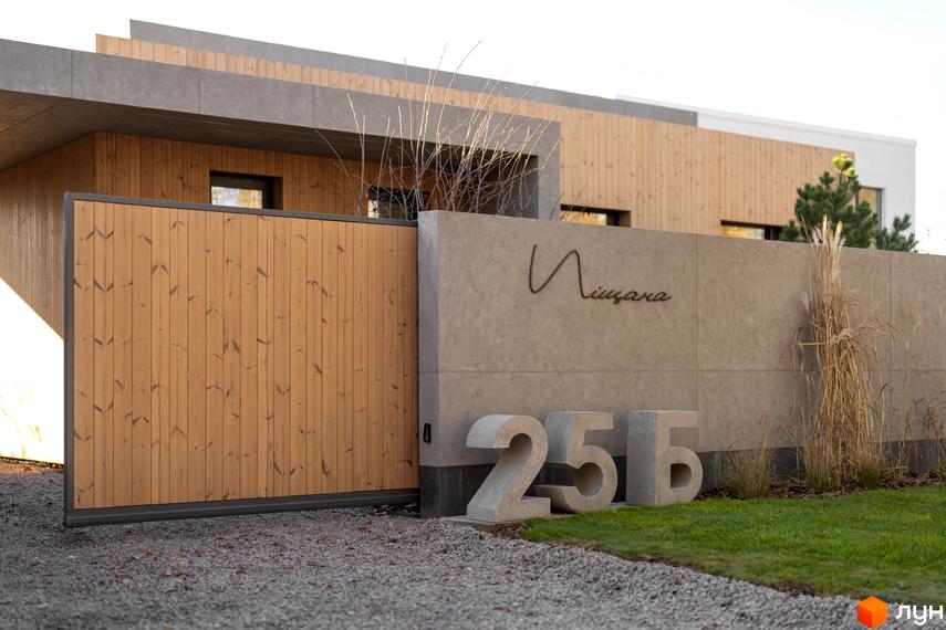 Житловий комплекс з дерев'яними фасадами та бетонним парканом, на якому вказано назву вулиці Піщана та номери будинків 25а, 25б. На території газон та гравій.