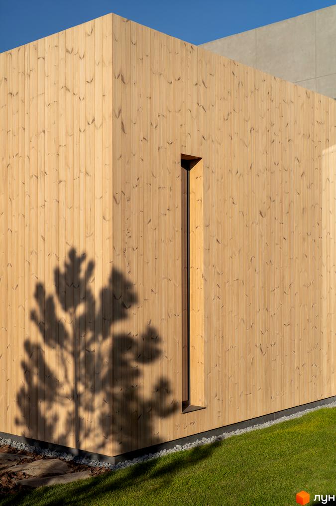 Світлий дерев'яний фасад будівлі з вузьким вікном, на тлі блакитного неба. На стіні тінь від гілок дерев, перед будинком газон та каміння.