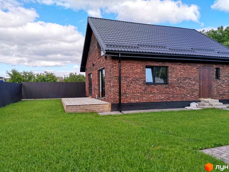 Котеджне містечко КМ Нова Баварія: цегляний будинок з дерев'яною терасою та доглянутим газоном. Навколо будинку – огорожа та зелені насадження.