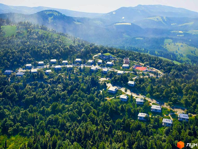 Котеджне містечко КМ GORA: білі будинки з сірими дахами, розташовані на схилі гори серед зелені, з видом на гірський пейзаж.