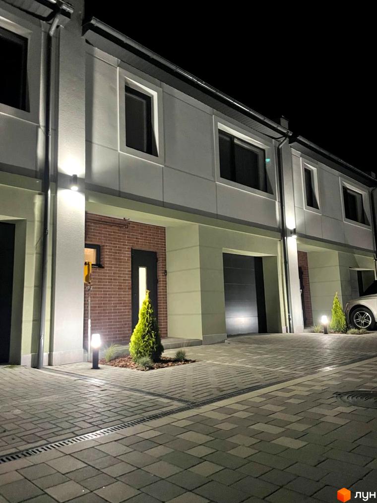 Таунхауси „Садовий Клуб“ з фасадом з сірої штукатурки та цегли, з гаражними воротами та підсвіткою. Прибудинкова територія з бруківки та зелених насаджень.