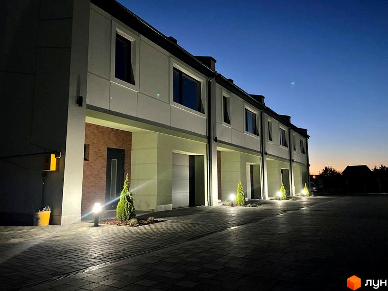 Таунхауси „Садовий Клуб“: фасади світлих будинків з темними вікнами та гаражами, освітлена прибудинкова територія з бруківкою та деревами.