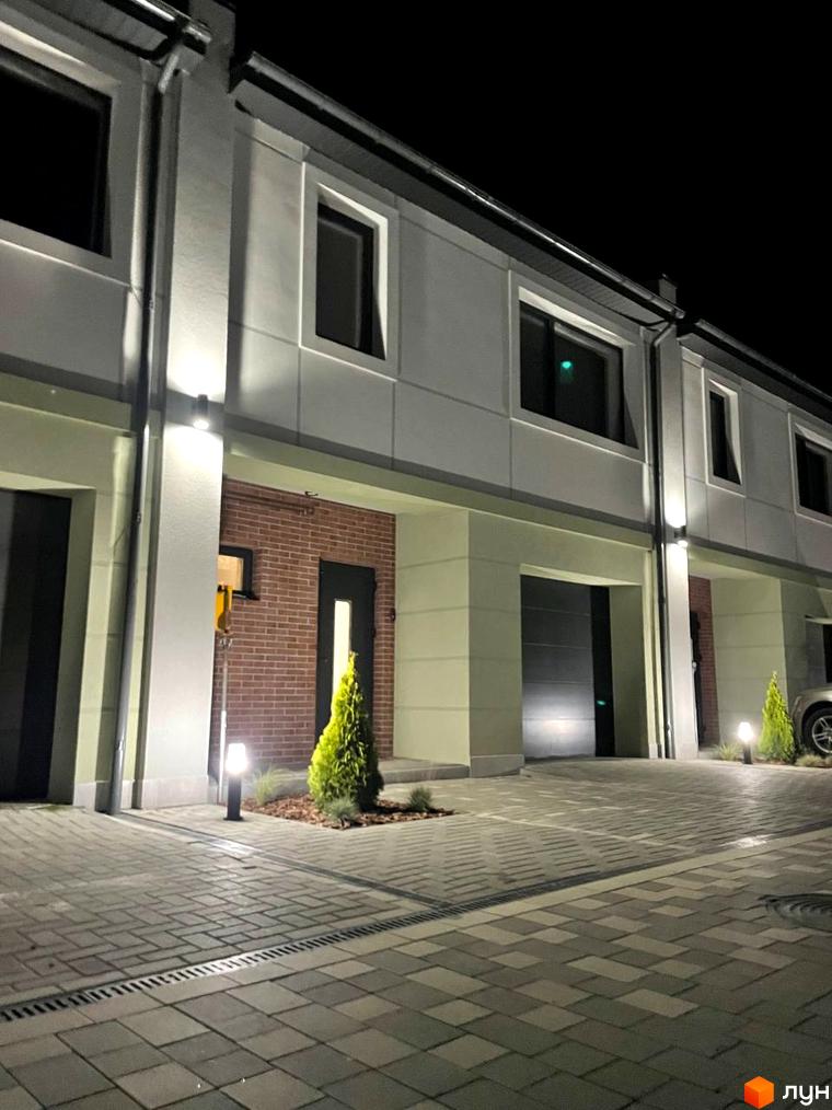 Таунхауси „Садовий Клуб“ з підсвіченими фасадами та гаражними воротами, вимощені бруківкою двір та зелені насадження.