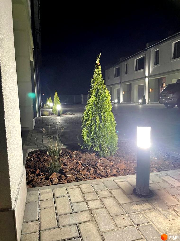 Таунхауси „Садовий Клуб“ з підсвіченою прибудинковою територією та елегантними ліхтарями. Сучасна архітектура новобудов та доглянутий ландшафт створюють затишну атмосферу.