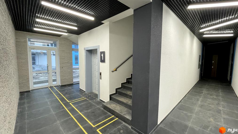 Вхідна група Житлового комплексу One Family з ліфтом, сходами та темною плиткою на підлозі. Приміщення має світлі стіни та чорну ґратчасту стелю з освітленням.