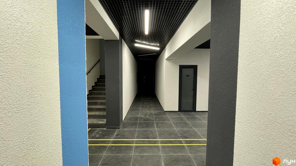 Внутрішній коридор Житлового комплексу One Family зі сходами, сітчастою стелею, світлими текстурованими стінами та темною плиткою.