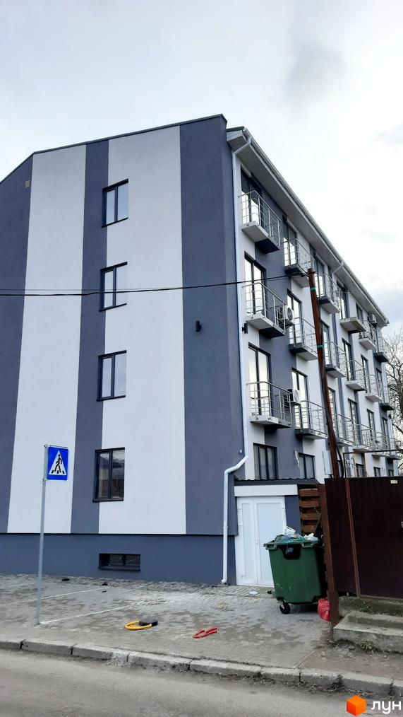 Фасад новобудови на вул. Осіння, 71, з сіро-білими смугами та балконами. Поруч розташована прибудинкова територія з пішохідним переходом та сміттєвими баками.