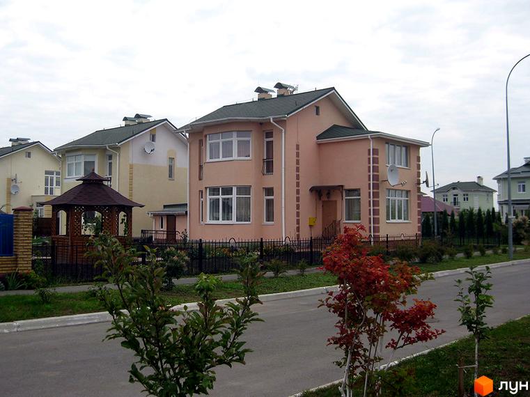 Двоповерховий будинок персикового кольору з білими вікнами. Поряд з будинком розташована доглянута прибудинкова територія з деревами та газоном.