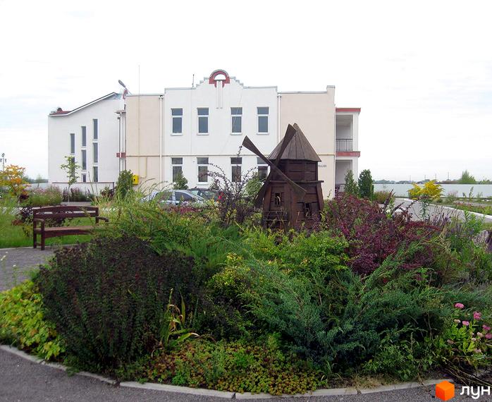 Житловий комплекс КМ Нова Богданівка. Біла будівля з вікнами та декоративним елементом на даху. На передньому плані ландшафтний дизайн з рослинами та дерев'яним млином.