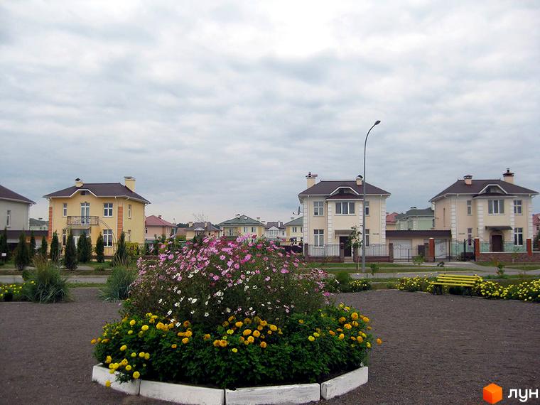 Двоповерховий будинок з коричневим дахом та білими стінами. На передньому плані клумба з квітами, навколо будинку доглянута територія.