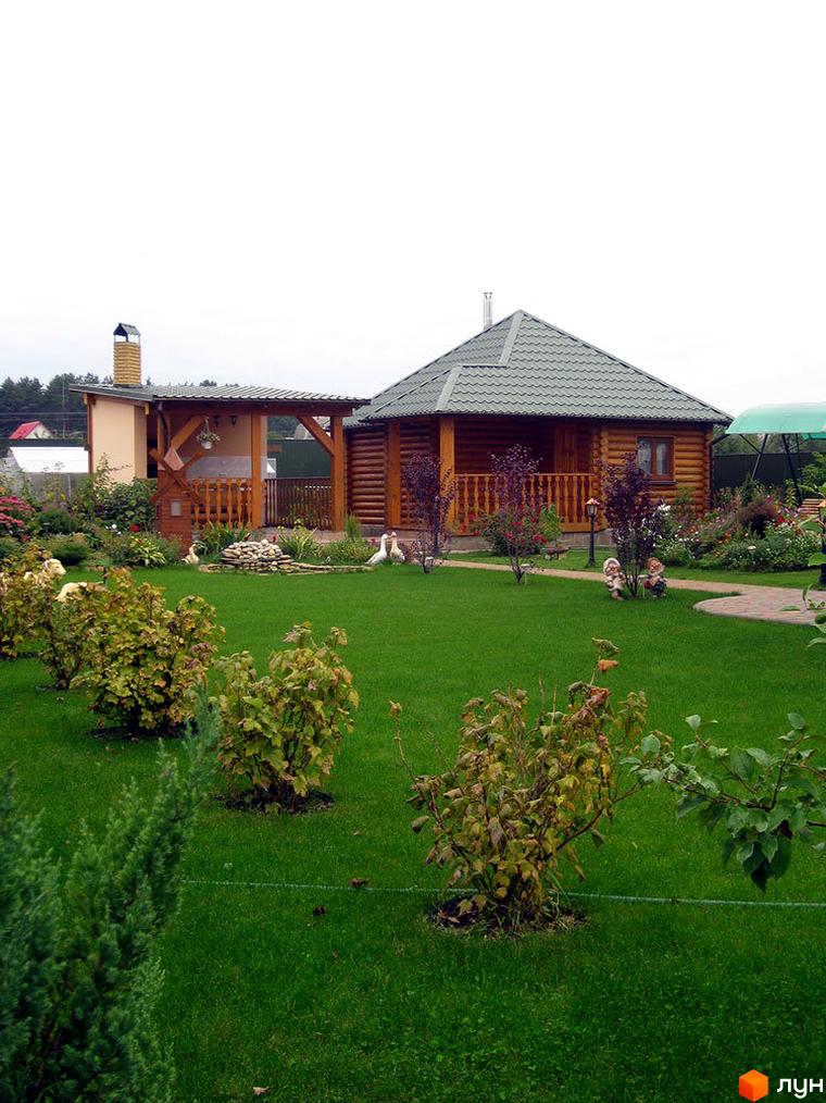 Дерев'яний будинок з зеленим дахом та терасою. На території розташовані газони, кущі та декоративні фігури.
