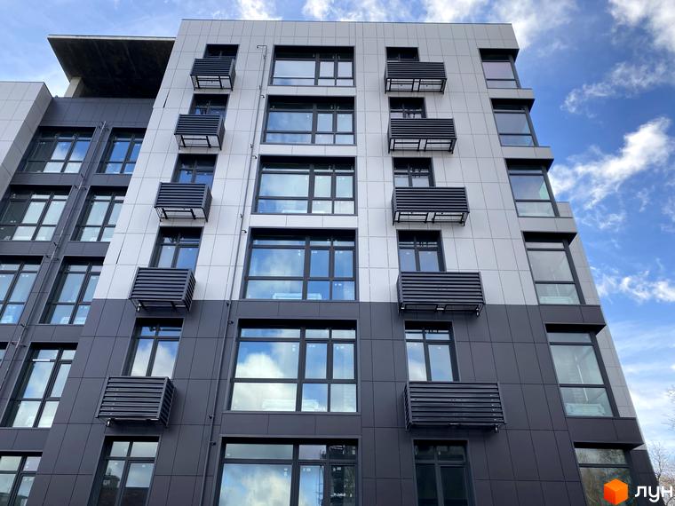 Зовнішній вигляд новобудови ЖК Forest Hill з сірими та білими фасадами, великими вікнами та кондиціонерами. Будинок розташований на фоні блакитного неба з хмарами.