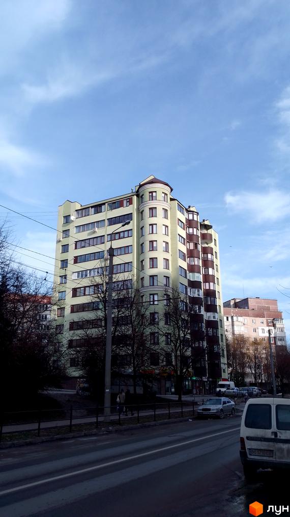 Фасад багатоповерхового житлового будинку світло-зеленого кольору з коричневими вікнами та округлою вежею. Поруч дорога, дерева та припарковані авто.