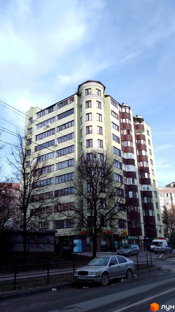 Багатоповерховий житловий будинок світлого відтінку з темними вікнами та округлою вежею. Навколо будинку є дерева, дорога та припарковані авто.