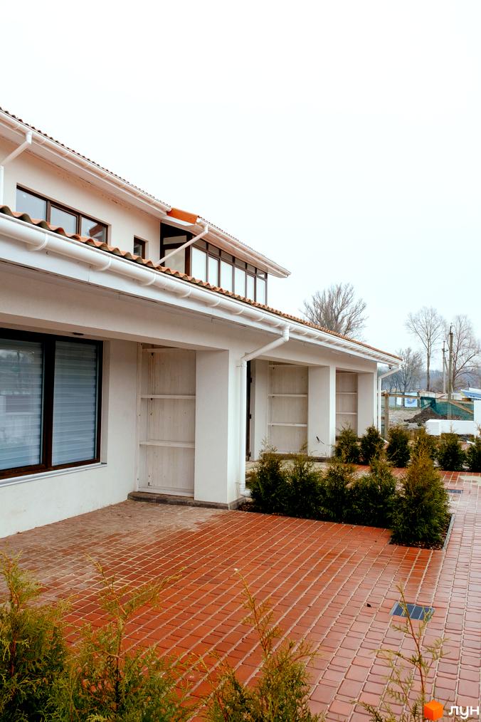 Зовнішній вигляд будинку КМ Little Italy Apartments з білою стіною, коричневим дахом та цегляною доріжкою. Прибудинкова територія озеленена туями.