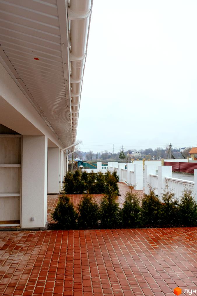 Зовнішній вигляд будівлі з білим фасадом та колонами, що веде до прибудинкової території з бруківкою та зеленими насадженнями житлового комплексу Little Italy Apartments.