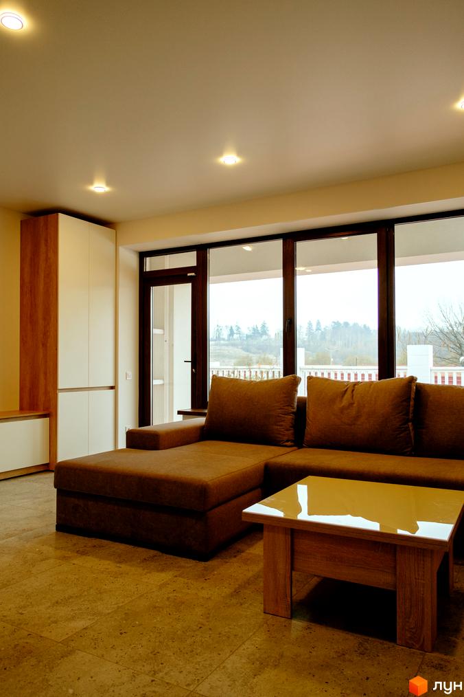 Інтер'єр вітальні в житловому комплексі КМ Little Italy Apartments з коричневим диваном та журнальним столиком. Великі вікна виходять на зелену територію.