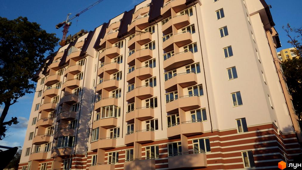 Житловий комплекс Будинок на Кузьмінській. Багатоповерховий будинок з балконами, оздоблений у світлих тонах з коричневими акцентами. На фоні - блакитне небо та будівельний кран.