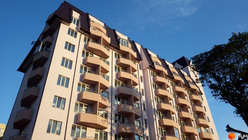 Житловий комплекс Будинок на Кузьмінській. Багатоповерховий будинок з балконами та дахом. Навколо будинку блакитне небо та дерева.