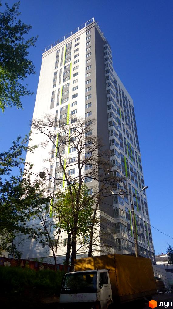 Висотна новобудова ЖК Лук'янівський з білими та сірими фасадами, зеленими акцентами та численними вікнами. На передньому плані видно дерева та вантажівку.